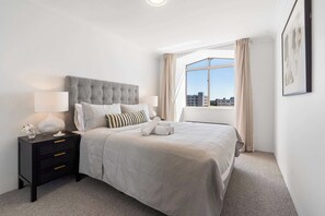 A neatly arranged hotel room with a large bed, a bedside table with a lamp, and a view of the city through a large window.