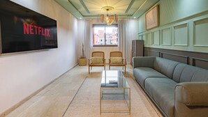 A living room with a flat-screen TV displaying 'NETFLIX', a sofa, two armchairs, a glass coffee table, and a view of buildings through a window. - Appartement de luxe au centre de Gueliz  (Marrakech)