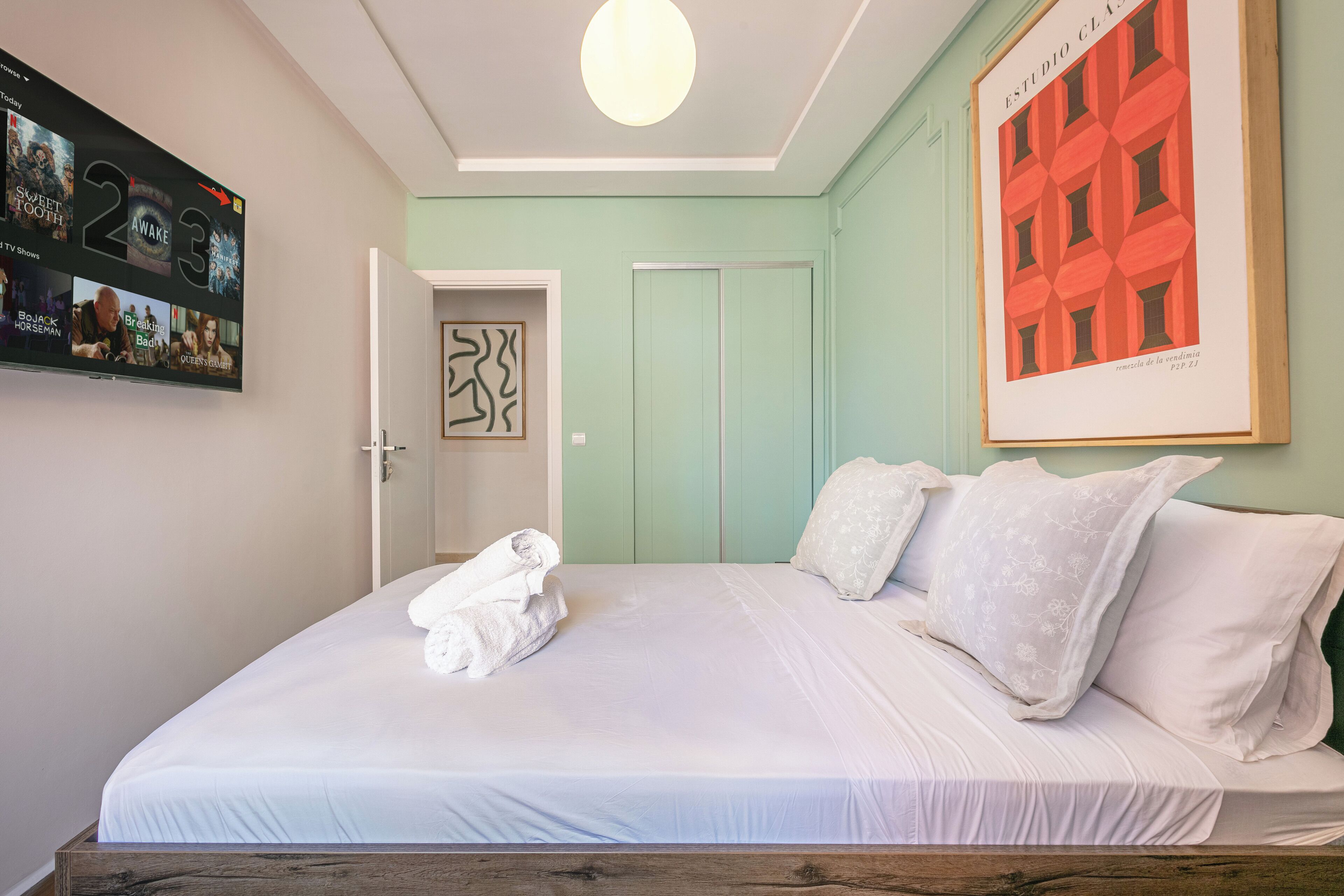 A neatly made bed with white linens and pillows, a framed artwork on the wall, and a TV displaying movie posters.