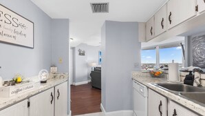 A modern kitchen with light wood cabinets, a marble countertop, and a view to the living area. - The Serenity at Peck Plaza (Daytona Beach)