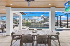 A modern outdoor dining area with a wooden table and wicker chairs, overlooking a swimming pool and a residential neighborhood. - Exquisite Waterfront Home | Featuring Pool/Hot Tub (Marco Island)