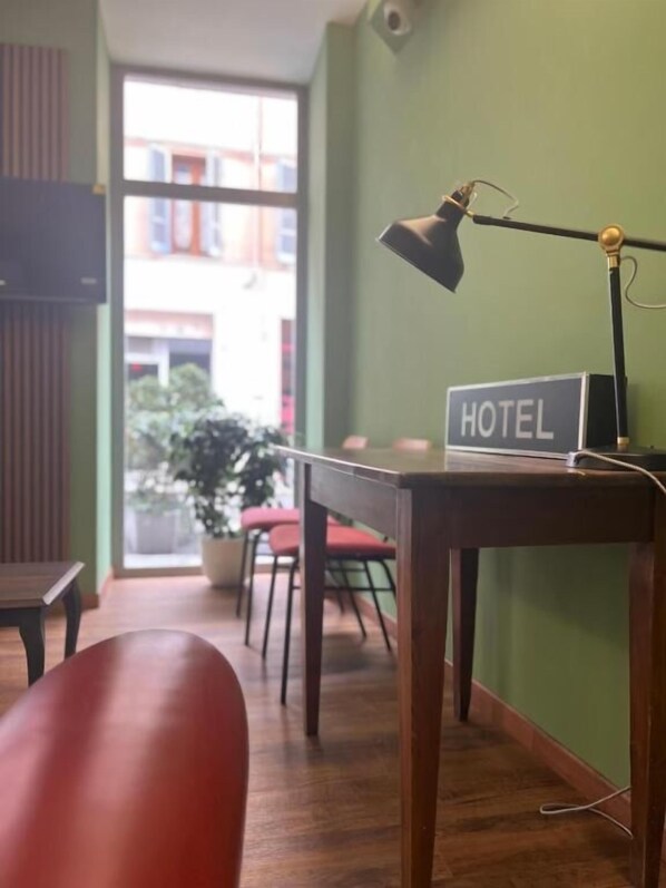 A hotel room with a wooden desk, a lamp, a sign reading "HOTEL", and a view of the outside through a window. - HOTEL BRENTA (Parma)