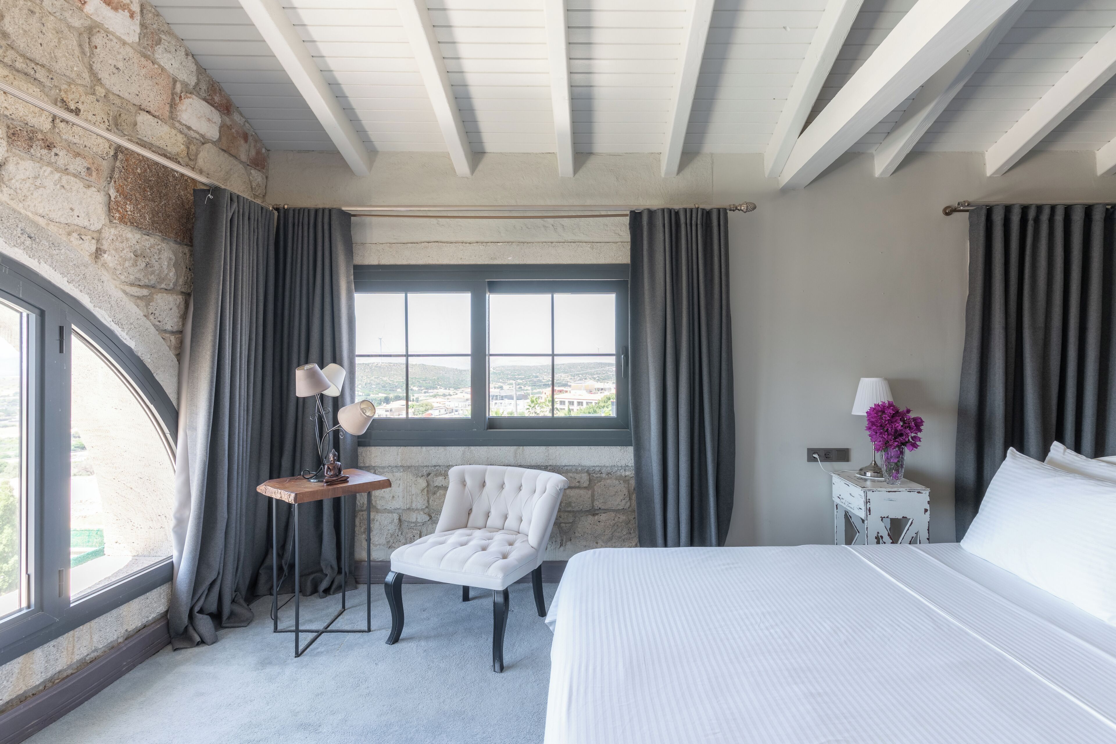 Ein Hotelzimmer mit einem großen Bett, einem kleinen Sessel, einem Nachttisch mit Lampe und Blick auf die Stadt durch ein Fenster.