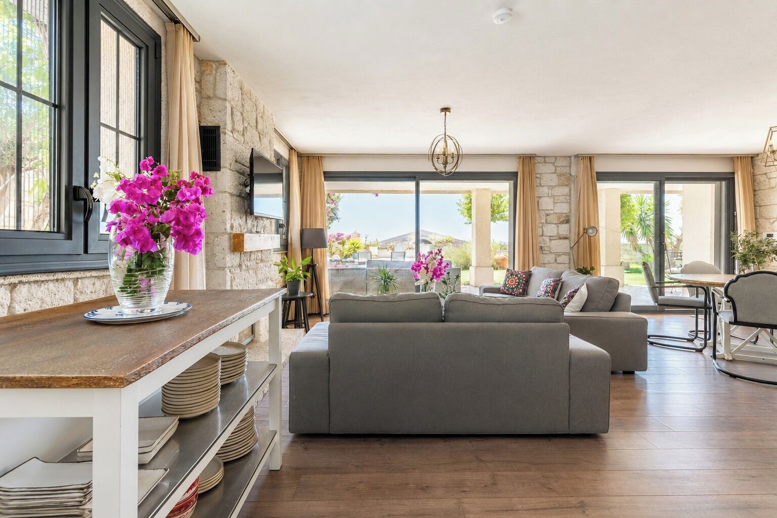 A spacious living area with a grey sofa, a wooden coffee table, and a vase of flowers. There is a stone wall and a large window providing a view outside.
