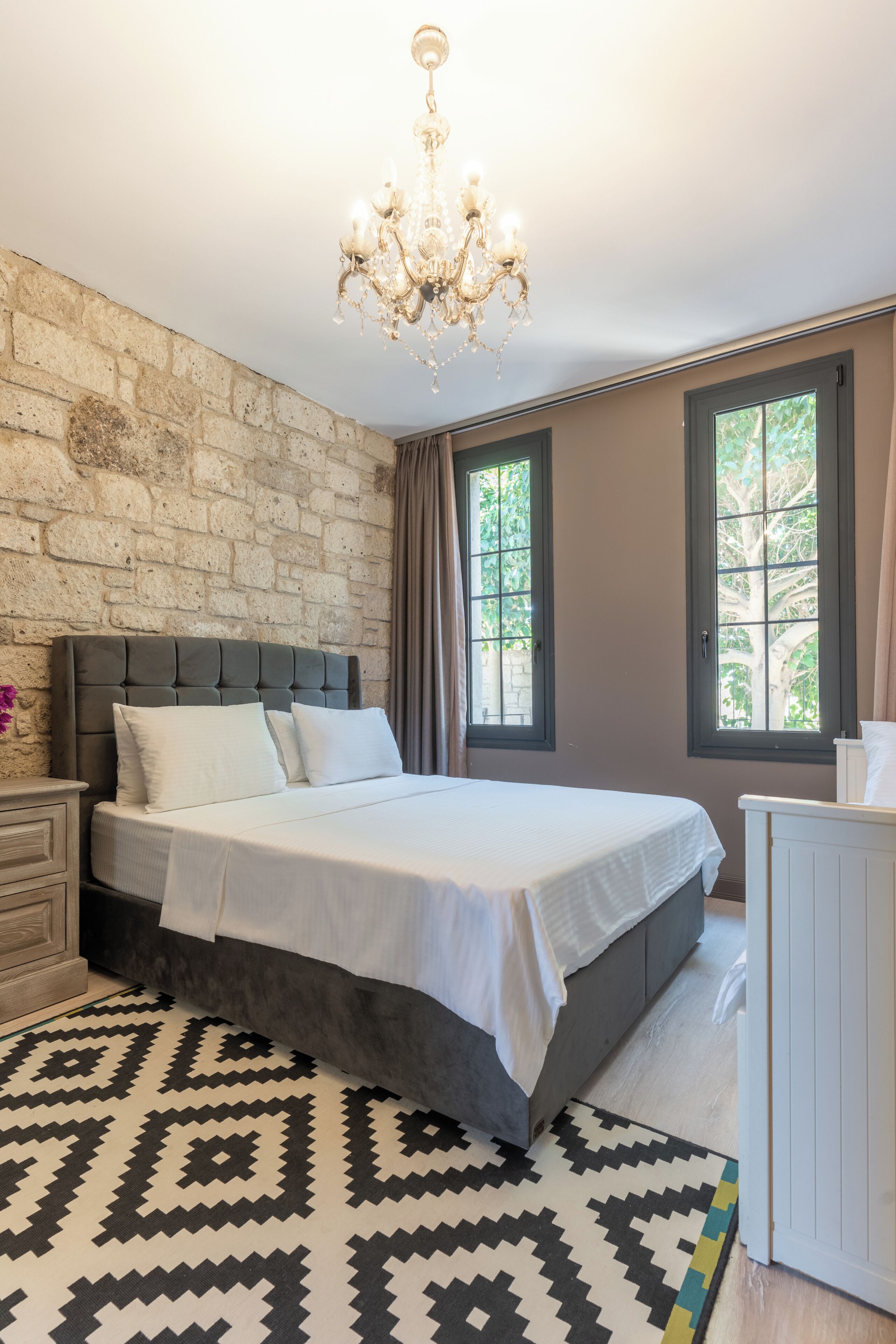 Een slaapkamer met een groot bed, een kroonluchter, een bakstenen muur en een raam met uitzicht op bomen.