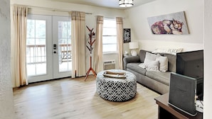 Un salon avec un canapé gris, un pouf rond, un téléviseur posé sur un meuble, un porte-manteau et un tableau accroché au mur.