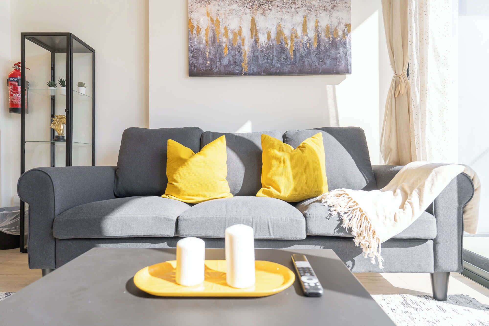 Una sala de estar moderna con un sofá gris, cojines amarillos, una mesa de centro negra con controles remotos y un cuadro en la pared.