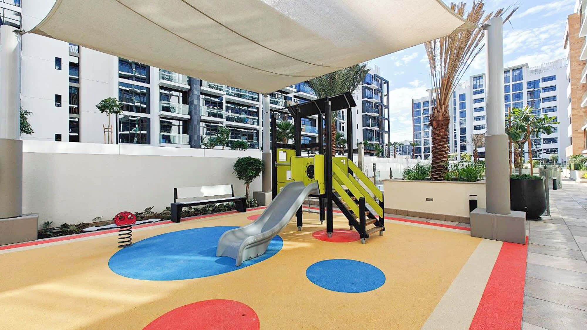 A playground with a slide and climbing structure, surrounded by a covered area and modern buildings in the background.