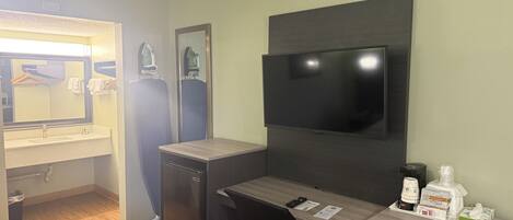 A compact hotel room with a kitchenette, a flat-screen TV mounted on the wall, a small desk with a chair, and a microwave.