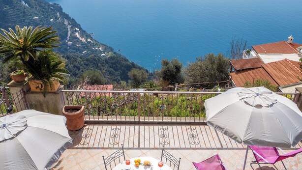 A terrace with a table set for dining, surrounded by potted plants and umbrellas, overlooking a coastal landscape with a mountain in the background. - Villa Lavinia (Positano)