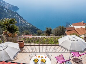 A terrace with a table set for dining, surrounded by potted plants and umbrellas, overlooking a coastal landscape with a mountain in the background.
