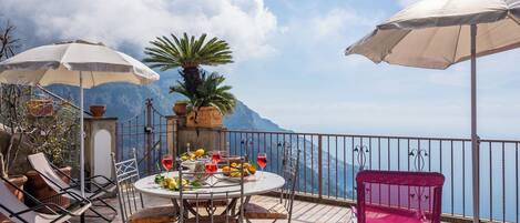 A terrace with a table set for two, surrounded by chairs and umbrellas, overlooking a mountainous landscape.