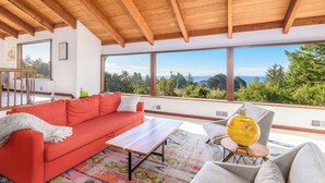 A modern living room with a red sofa, a coffee table, and a view of the forest and ocean through large windows. - Serene 3 BR Sleeps 8 with Hot Tub & EV Charger! (Sea Ranch)