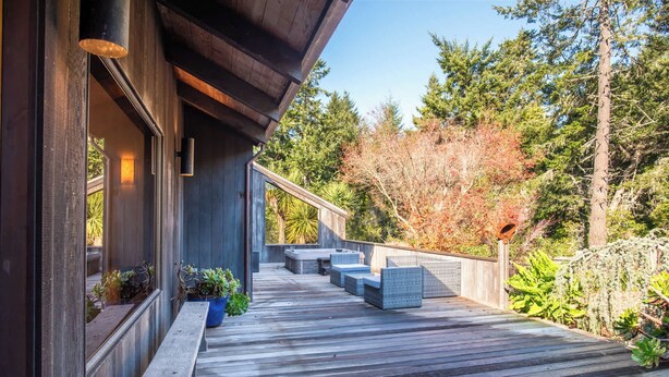A wooden deck with outdoor seating, surrounded by trees and vegetation. - Serene 3 BR Sleeps 8 with Hot Tub & EV Charger! (Sea Ranch)