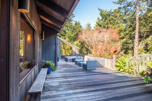 A wooden deck with outdoor seating, surrounded by trees and vegetation. - Serene 3 BR Sleeps 8 with Hot Tub & EV Charger! (Sea Ranch)