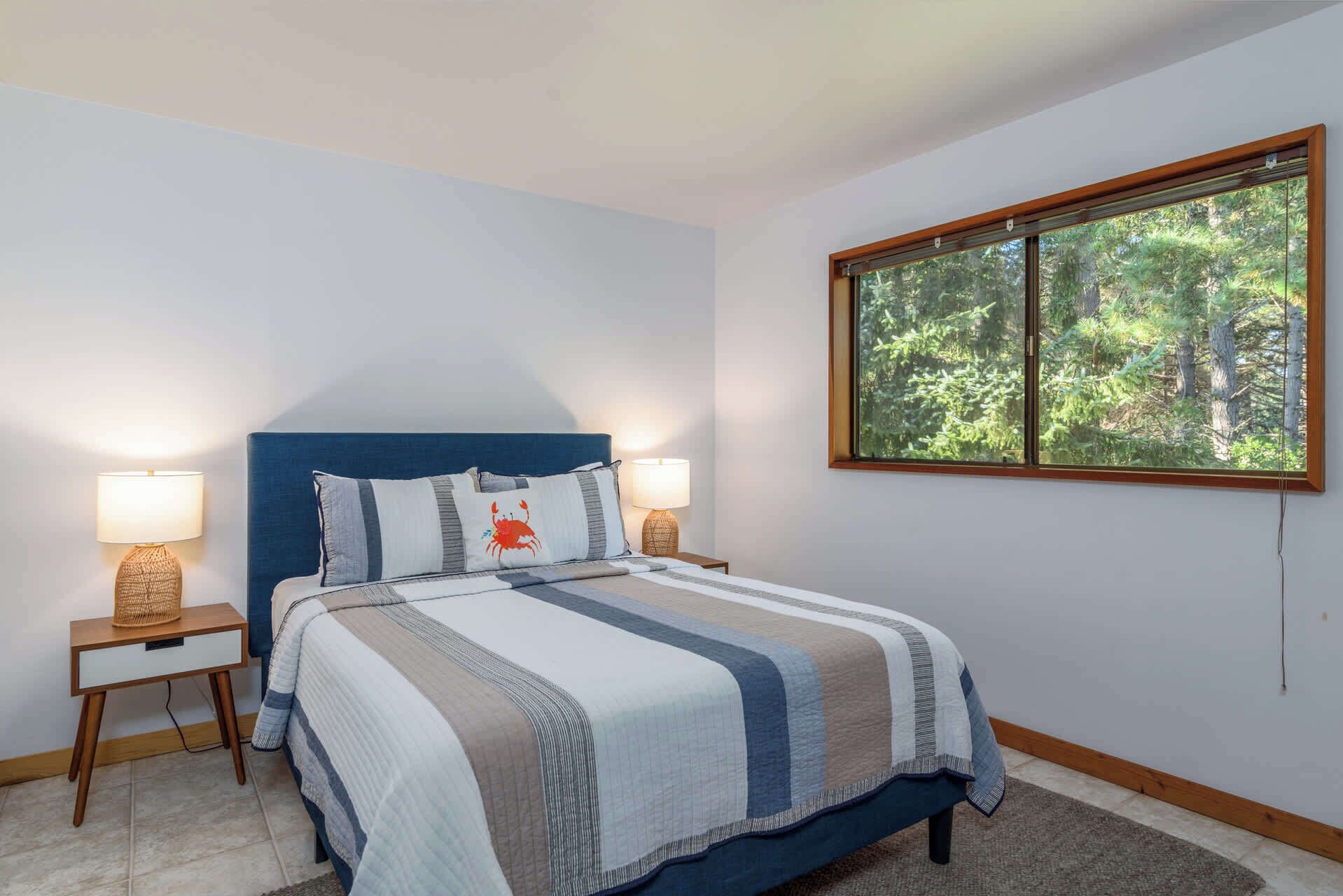 A bedroom with a bed, two bedside lamps, a window with a view of trees, and a blue headboard.