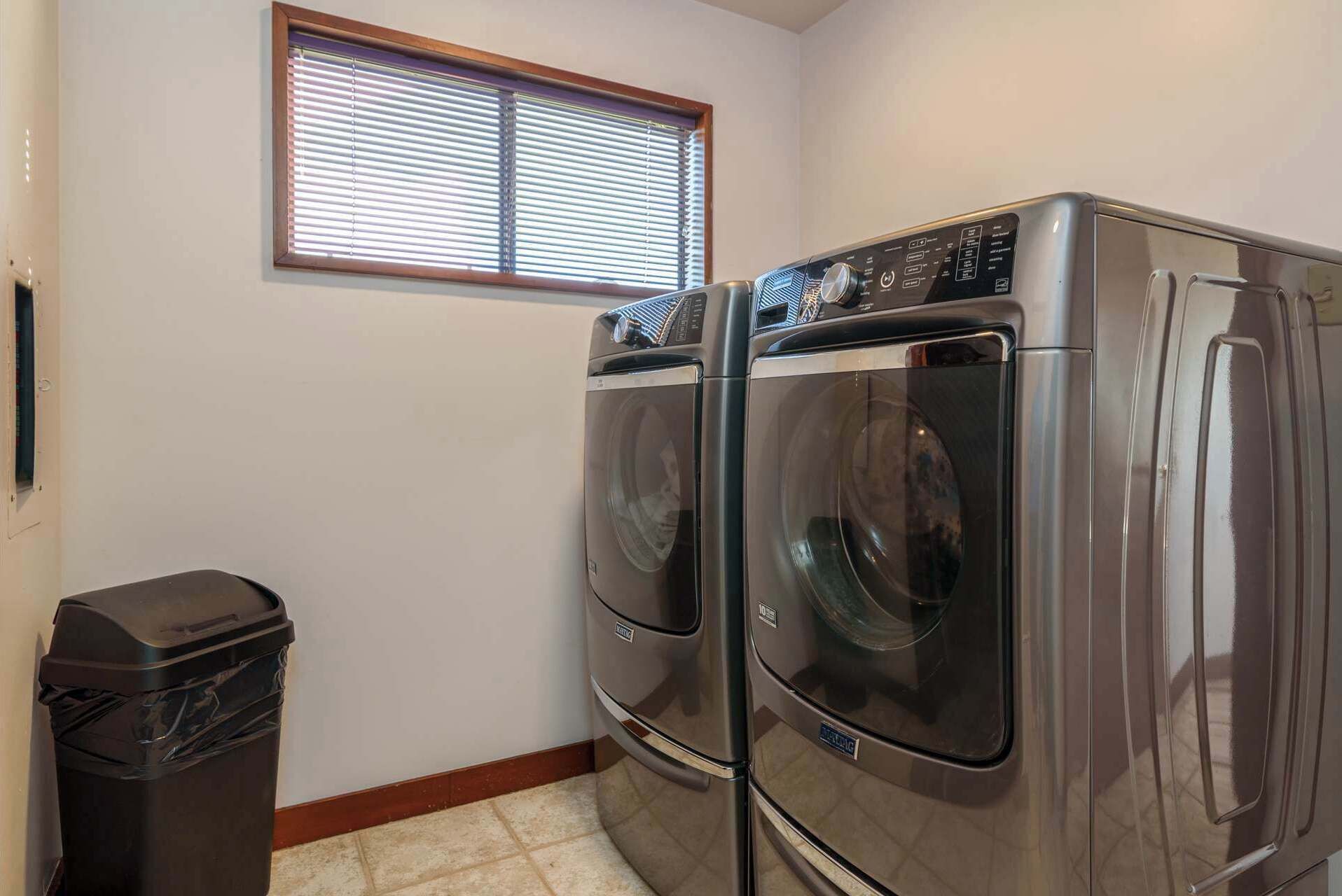 A laundry room with two front-loading washing machines and a trash can.