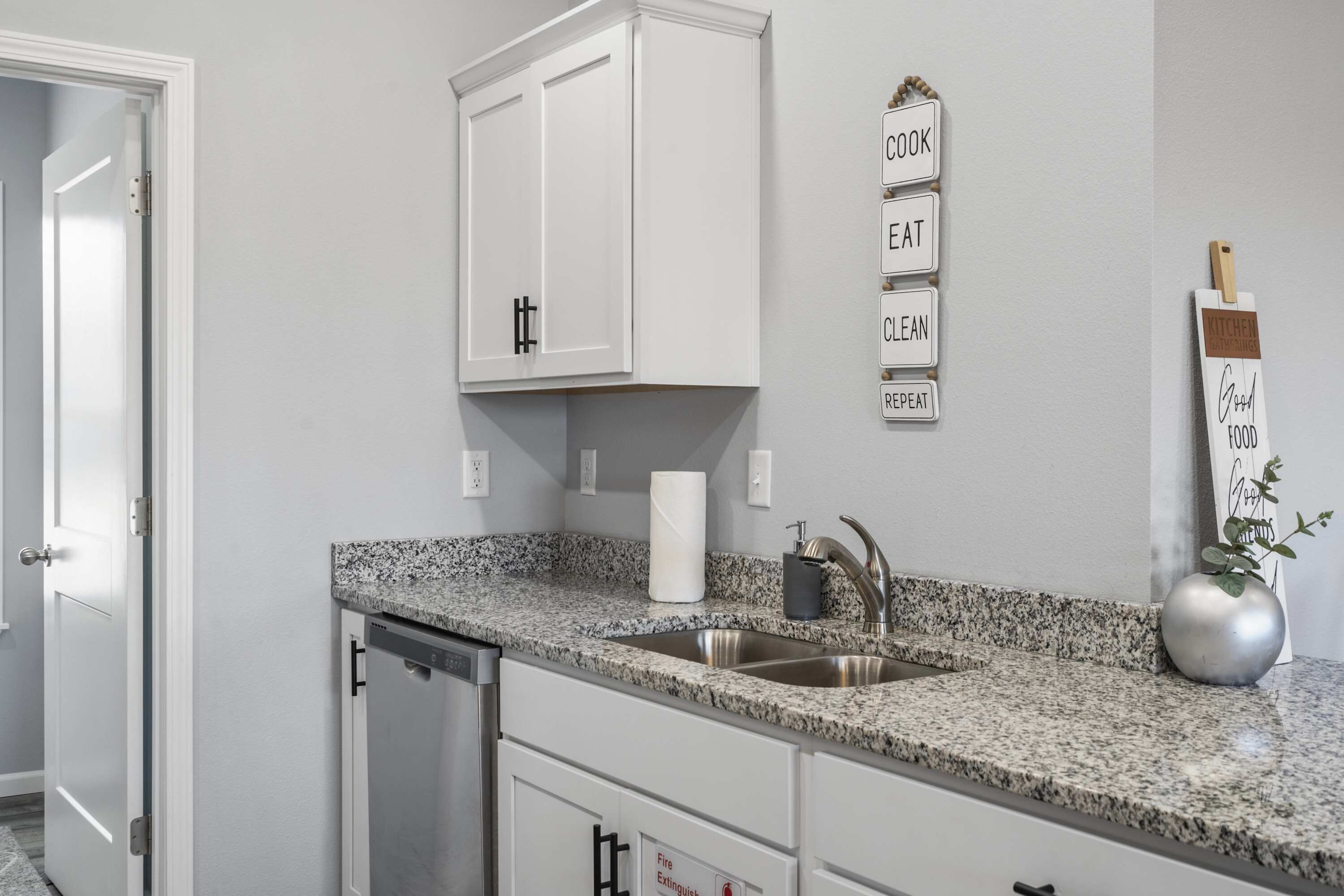 Una cocina con armarios blancos, encimera de granito, pileta de acero inoxidable, extintor de incendios y un cartel en la pared con términos relacionados a la cocina.