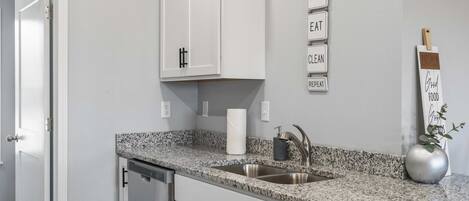 Una cocina con armarios blancos, encimera de granito, pileta de acero inoxidable, extintor de incendios y un cartel en la pared con términos relacionados a la cocina.