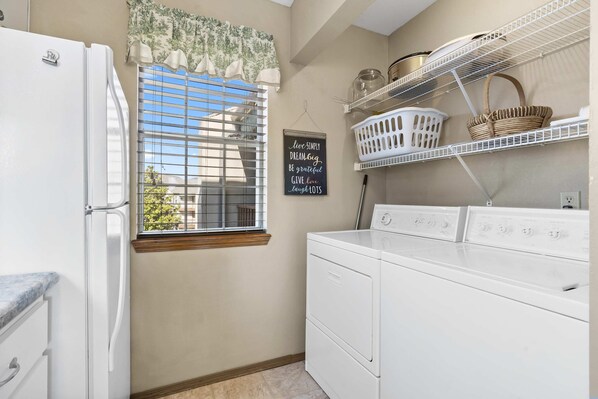 A laundry area with a washer and dryer, a window with blinds, a shelf with baskets, and a motivational quote on the wall.