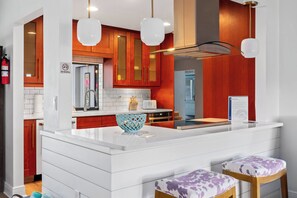 A modern kitchen with white countertops, a central island, and wooden cabinets. There are two bar stools with purple cushions.
