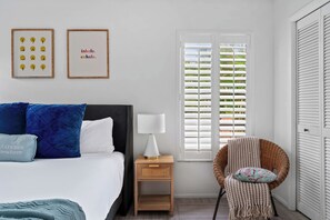 A bedroom with a bed, a nightstand with a lamp, a wicker chair, and a window with shutters.