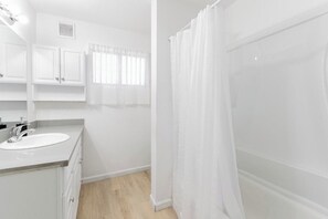 Un baño con lavamanos blanco, armarios, cortina de ducha y una ventana con cortinas.