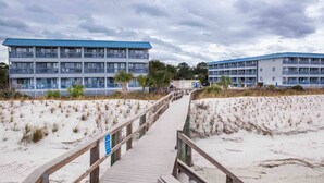 Une promenade en bois mène aux bâtiments situés en bord de mer, dotés de balcons, et l’ensemble est entouré de dunes de sable.