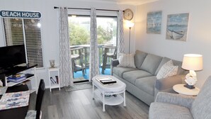 Un salon avec un canapé gris, une table basse blanche, un téléviseur et un panneau indiquant « MAISON DE PLAGE ».