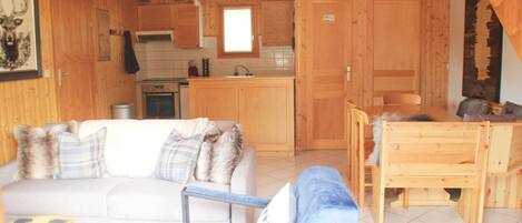 A wooden cabin interior with a living area featuring a sofa and armchairs, a dining table, and a kitchen with wooden cabinets and a window.