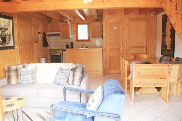 A wooden cabin interior with a living area featuring a sofa and armchairs, a dining table, and a kitchen with wooden cabinets and a window. - Chalet in La Béchigne with Valley Views (Châtel)