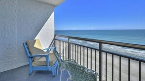 A balcony with a blue chair and a wicker chair, overlooking a beach with the ocean and a clear blue sky.