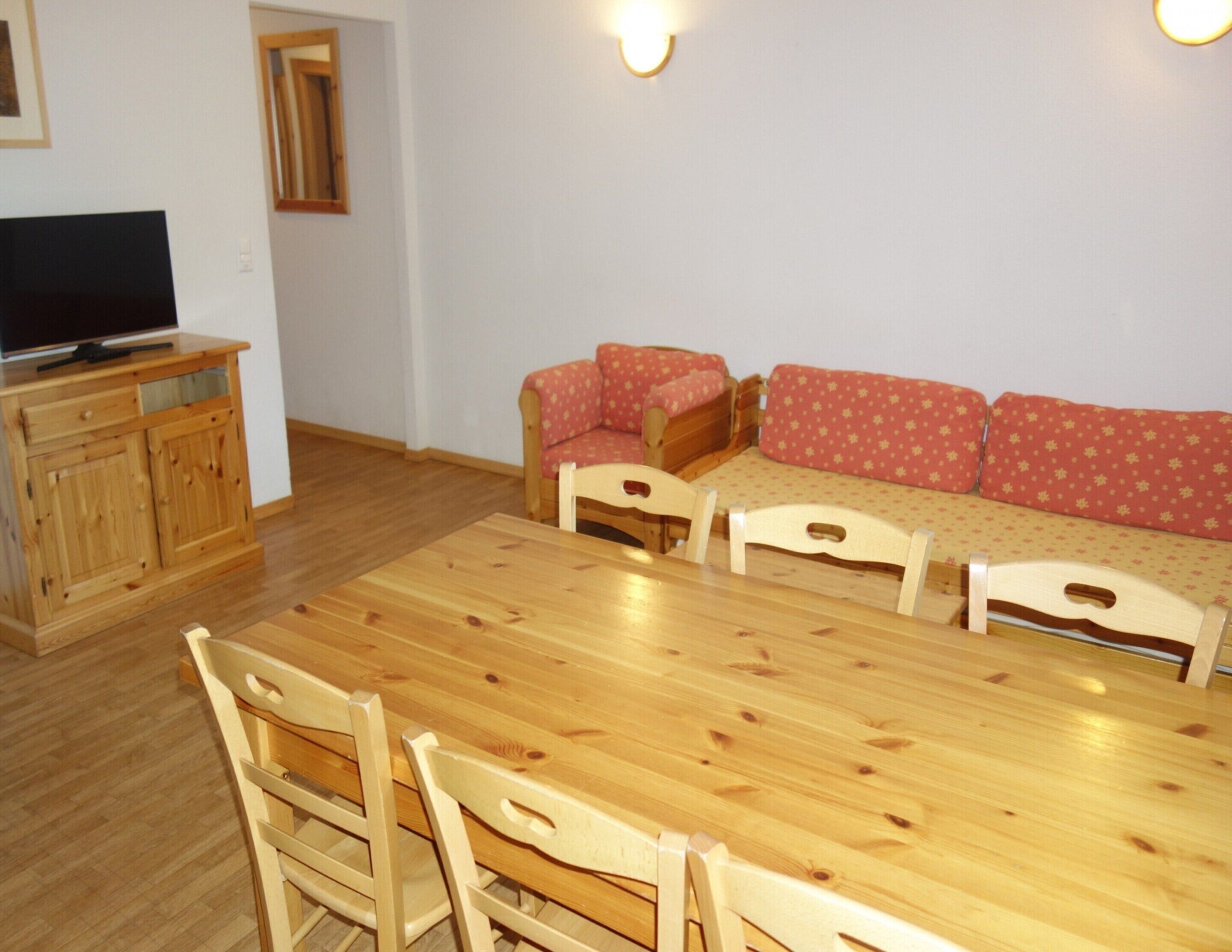 Uma sala de jantar com uma mesa e cadeiras de madeira, um conjunto de sofás e uma televisão sobre um armário de madeira.