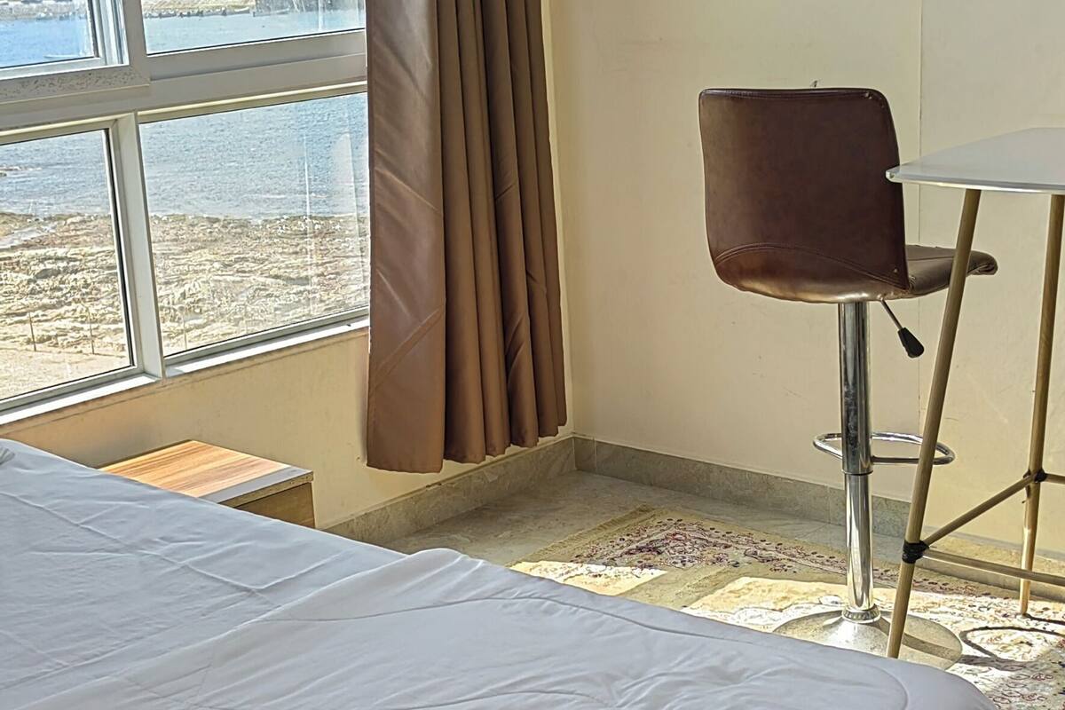 A hotel room with a large bed, a desk, and a chair. There are nautical-themed decorations on the walls, and a view of the sea through a large window.