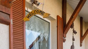 A window with a 'WELCOME' sign, wooden shutters, a hanging bell, and a potted plant.