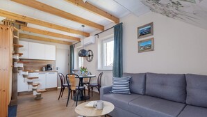 A modern living room with a grey sofa, wooden beams, a dining area with a table and chairs, and a kitchen in the background.