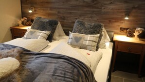 A room with two beds, each with a grey blanket and a pillow with a heart design. There are two bedside tables with decorative items and a wall with a wooden panel design. - Apartment Haus Tauern Am See  (Heiterwang)