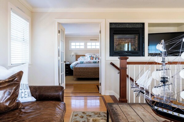 A living room with a brown leather sofa, a wooden coffee table, and a large ship model. There is a bed with a floral bedspread in the background.