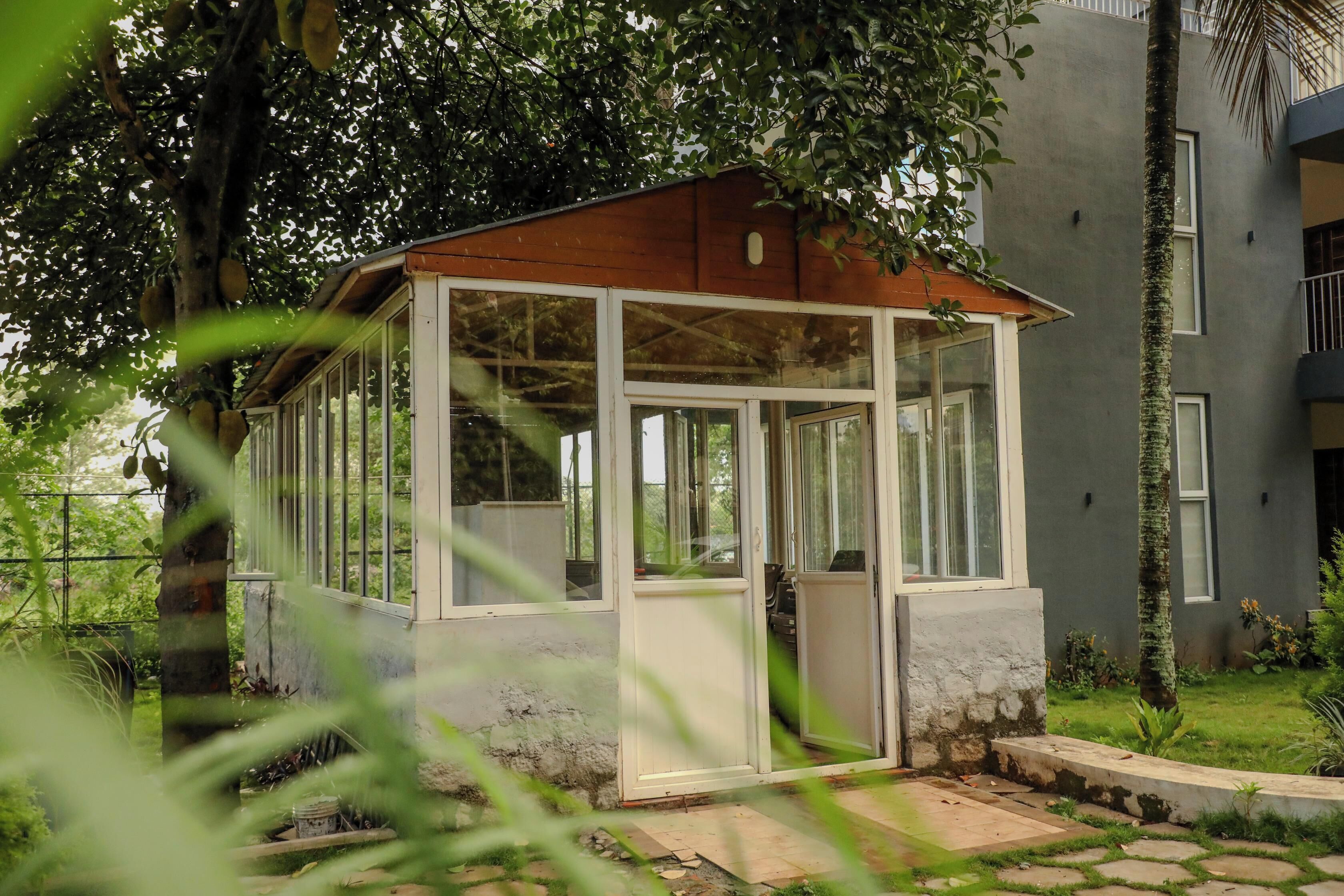 Una pequeña estructura cerrada con paredes de vidrio y techo de madera, rodeada de vegetación y un edificio al fondo.