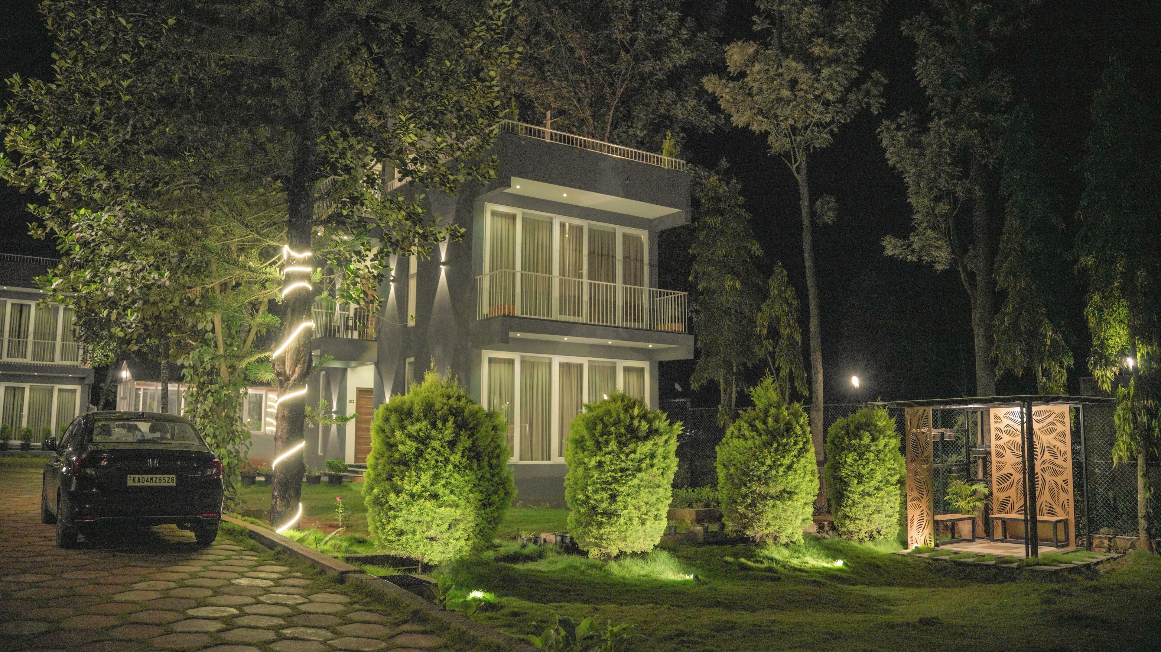A modern two-story building with a balcony, surrounded by lush greenery and a paved driveway.