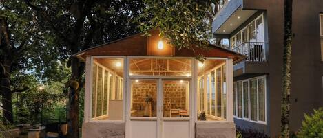 A cozy, well-lit wooden gazebo with a stone base, surrounded by lush greenery and a stone pathway.