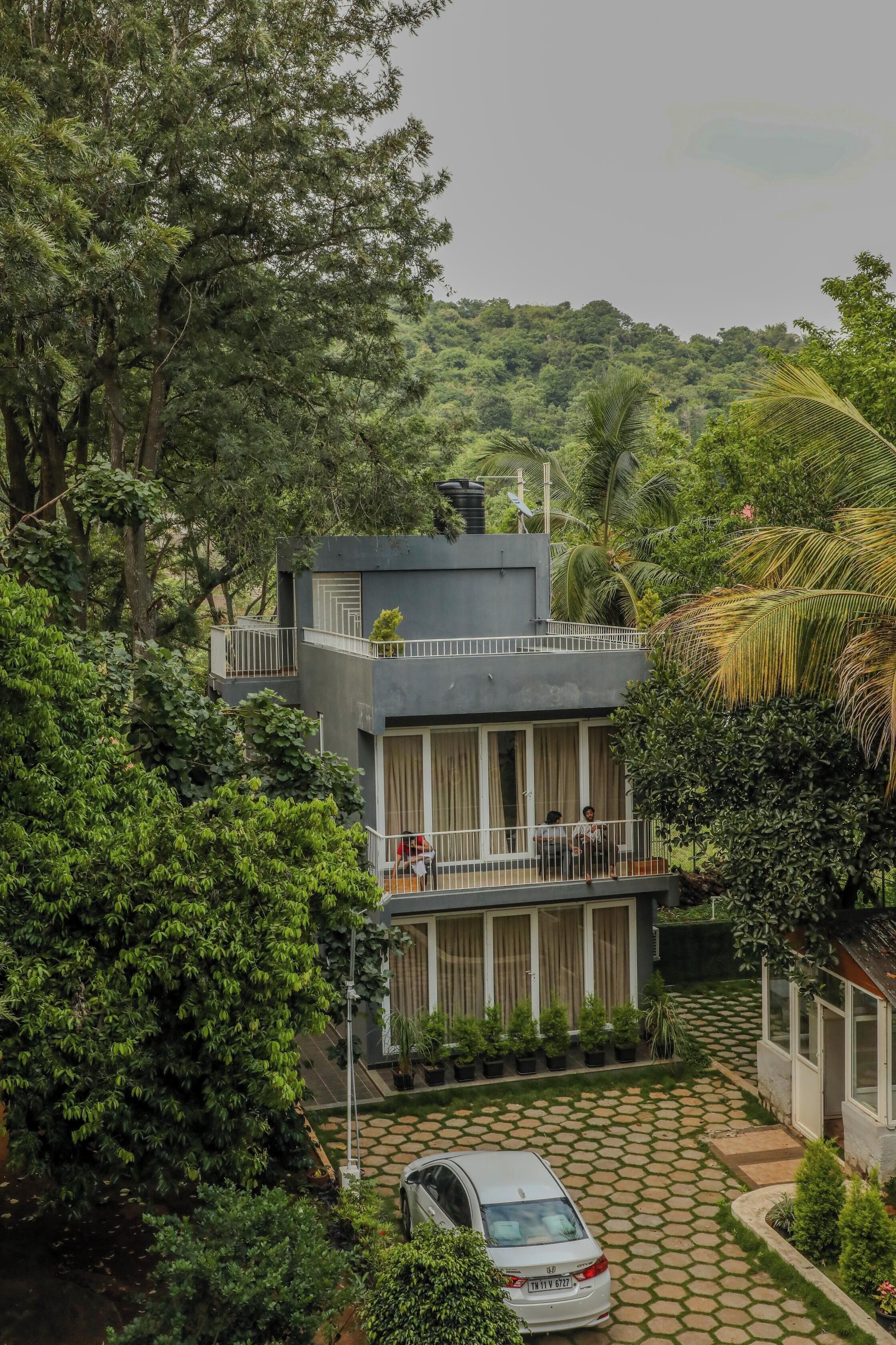 A modern two-story building with a balcony, surrounded by lush greenery and a hilly landscape.
