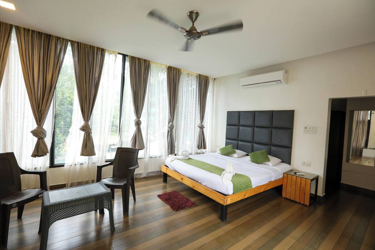 Quarto com cama, ventoinha no teto, janela com cortinas, mesa pequena e cadeira.