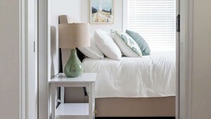 A bedroom with a bed, a nightstand with a lamp, a framed picture on the wall, and a window with blinds.