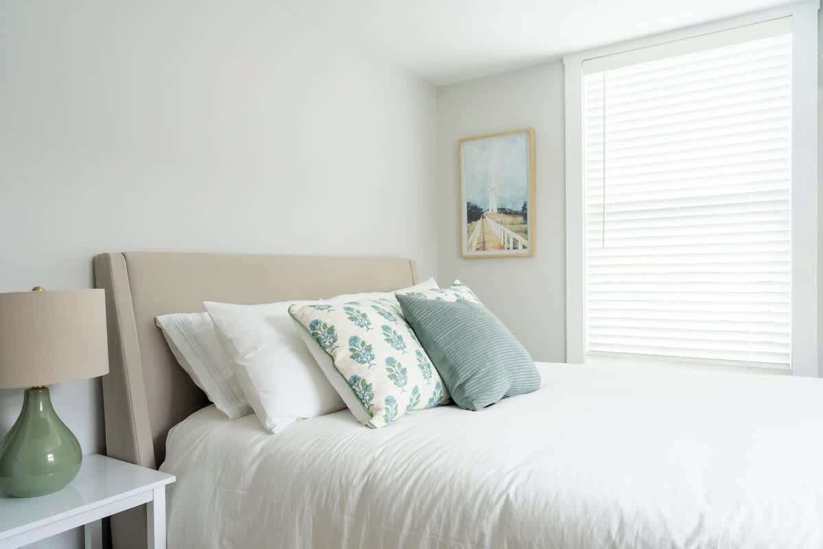 A bedroom with a bed, pillows, a nightstand, a lamp, a painting, and a window with blinds.