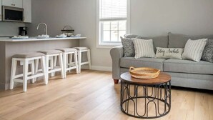 A modern kitchen with white cabinets, a microwave, and a coffee maker. There is a grey sofa with striped pillows and a basket on a round table.