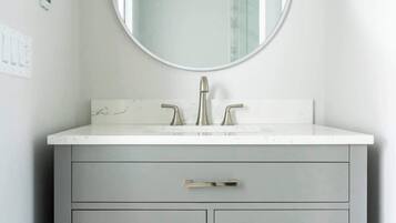 A bathroom with a round mirror, a white countertop, a single sink, and grey cabinets with drawers.