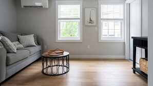 A living room with a grey sofa, two windows with blinds, a round table with a basket, and a black console table with a basket.