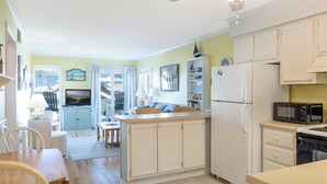 A compact kitchen with white cabinets, a refrigerator, a microwave, and a dining area with a table and chairs. - SeaSpray 350, Oceanview Condo in Atlantic Beach, NC! (Atlantic Beach)