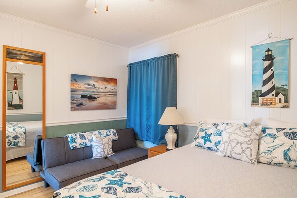 Un dormitorio con cama, sofá, lámpara, un cuadro de un faro y una ventana con cortinas azules.